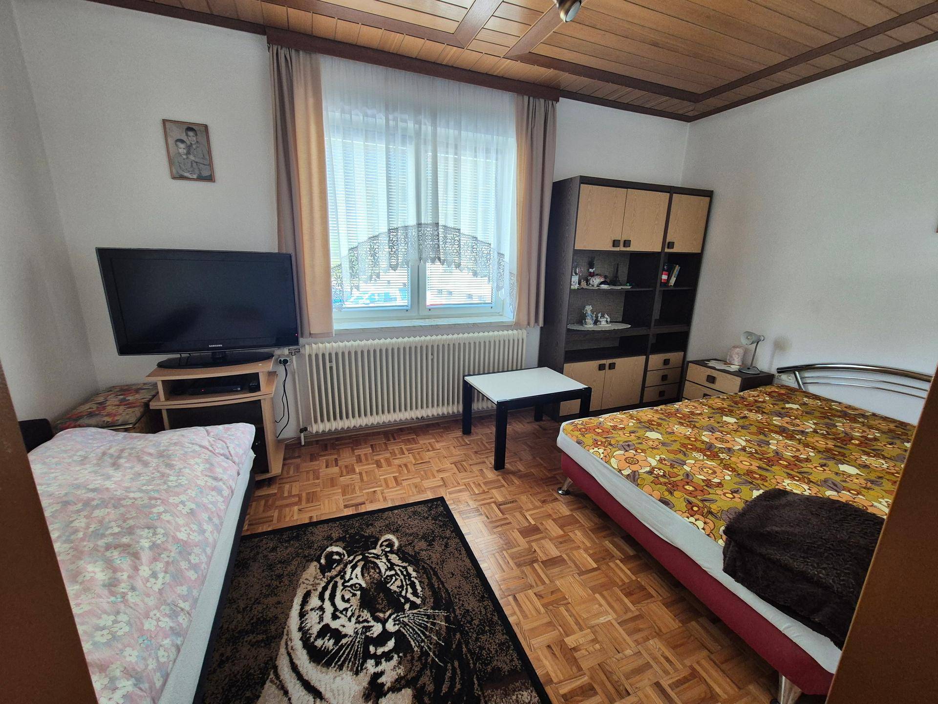 Schlafzimmer mit Parkettboden, Holzdecke und Fenster, eingerichtet mit Bett und Schrank.