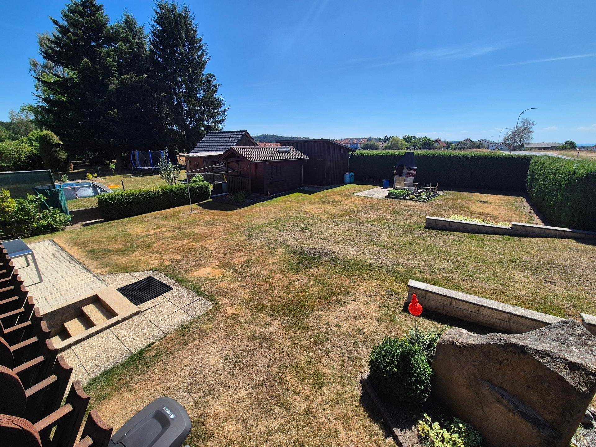 Großer Garten mit Rasenfläche, Hecken, Gartenhäusern und einem Grillplatz unter blauem Himmel.