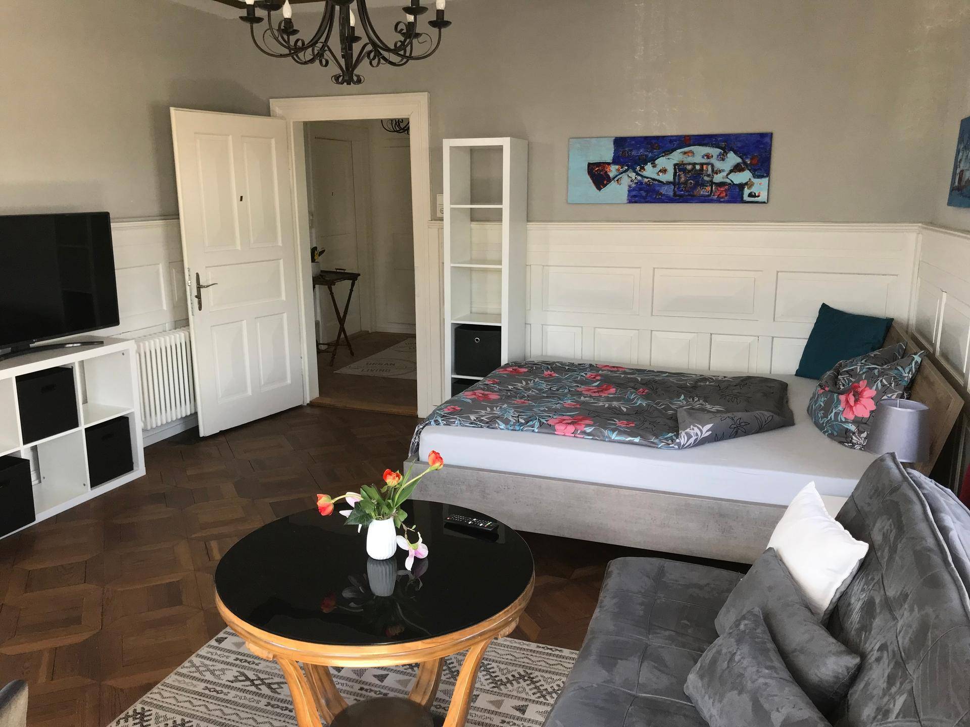 Gemütlicher Wohnbereich mit Parkettboden, Bett und Fernseher, durch eine Tür mit einem weiteren Raum verbunden.