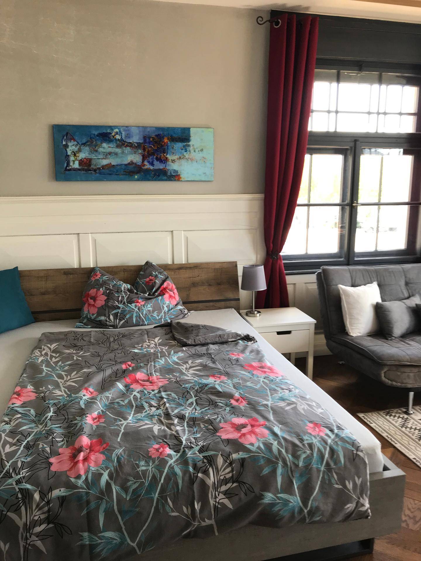 Gemütliches Schlafzimmer mit floraler Bettwäsche, modernem Kunstwerk und viel Tageslicht durch die Fenster.