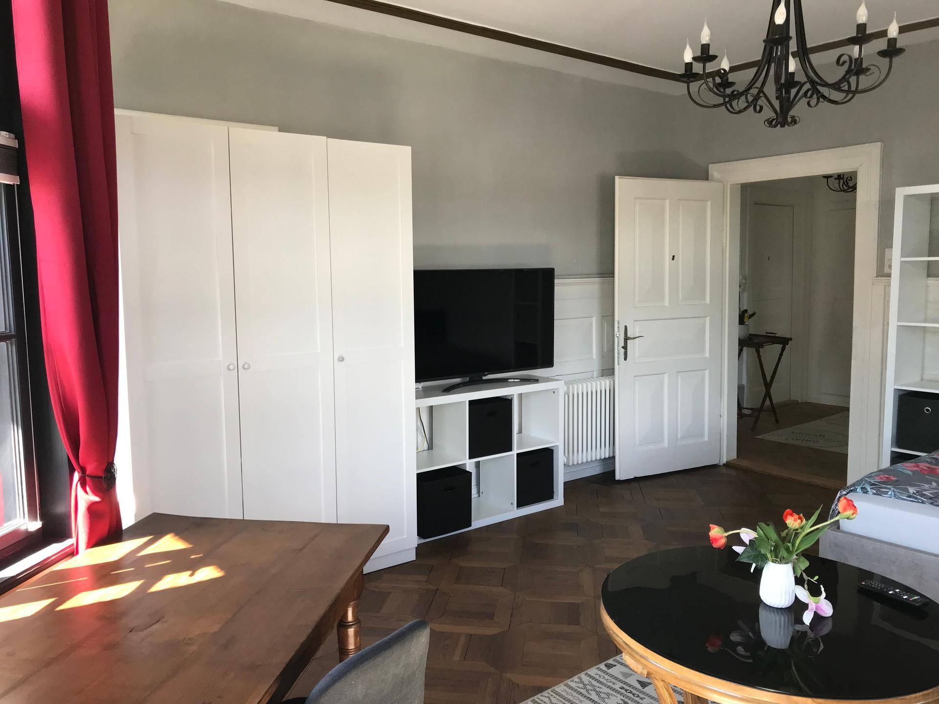 Heller Wohnbereich mit Fernseher, weißem Kleiderschrank und Zugang zu einem weiteren Raum.