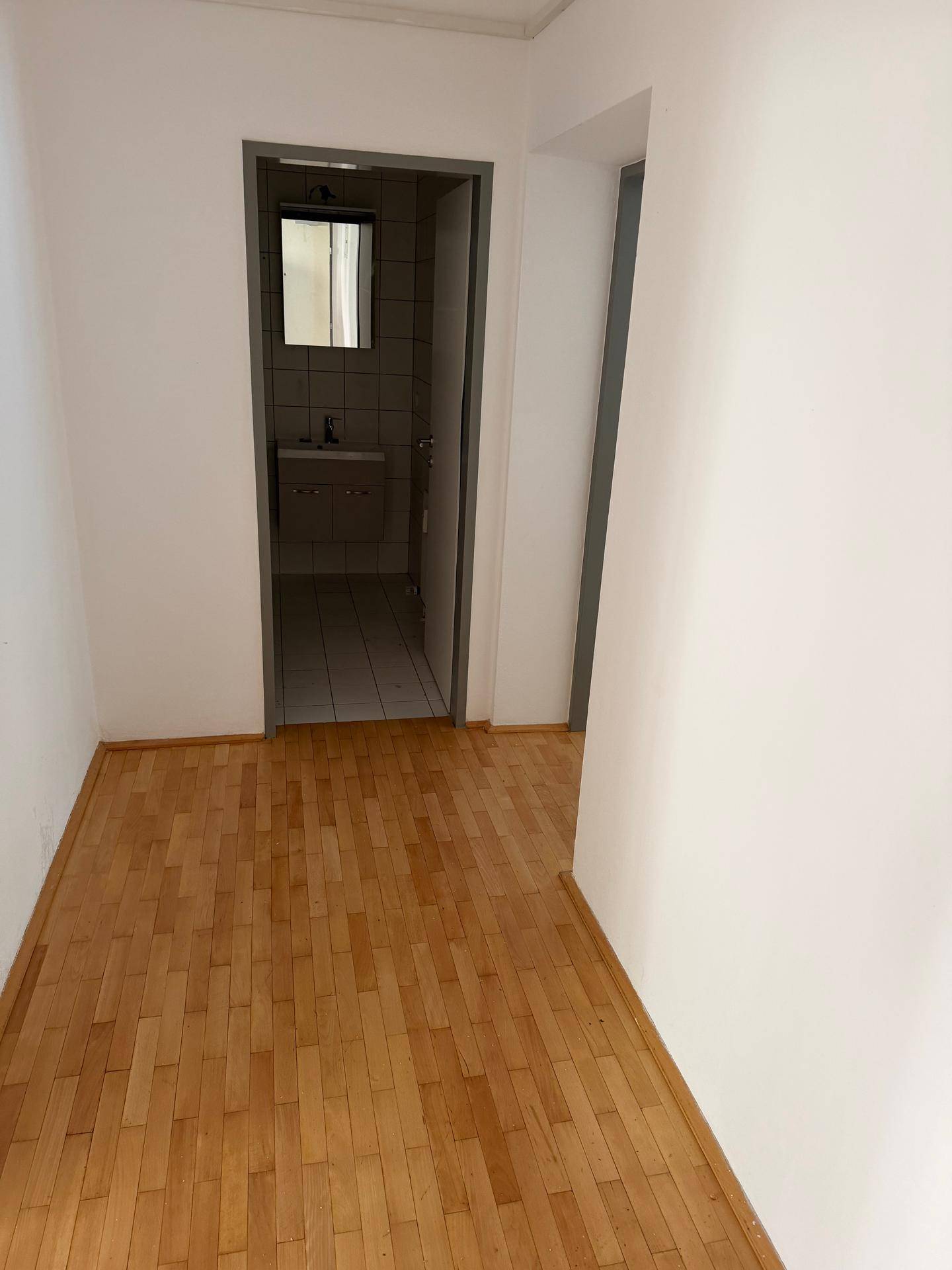 Heller Flur mit Parkettboden und weiß gestrichenen Wänden, der zum Badezimmer führt.