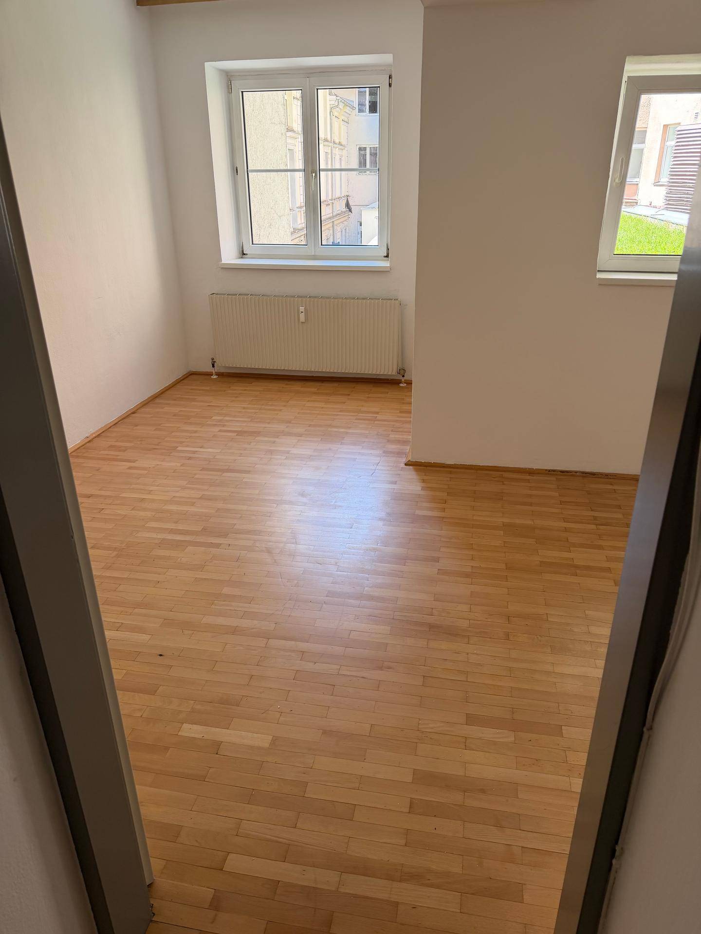 Leeres Zimmer mit hellem Parkettboden, weißen Wänden und einem Fenster mit Blick auf die Stadt.