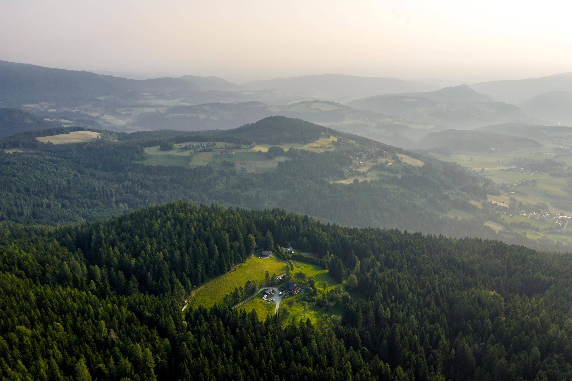 Luftaufnahme einer idyllischen Hügellandschaft mit dichten Wäldern, Wiesen und verstreuten Häusern.