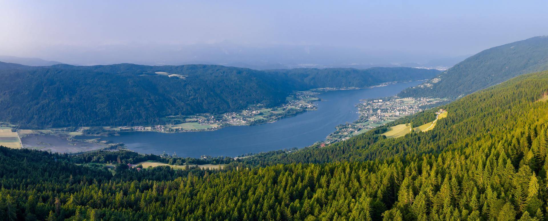 Atemberaubender Panoramablick auf einen See, umgeben von bewaldeten Bergen und kleinen Ortschaften.