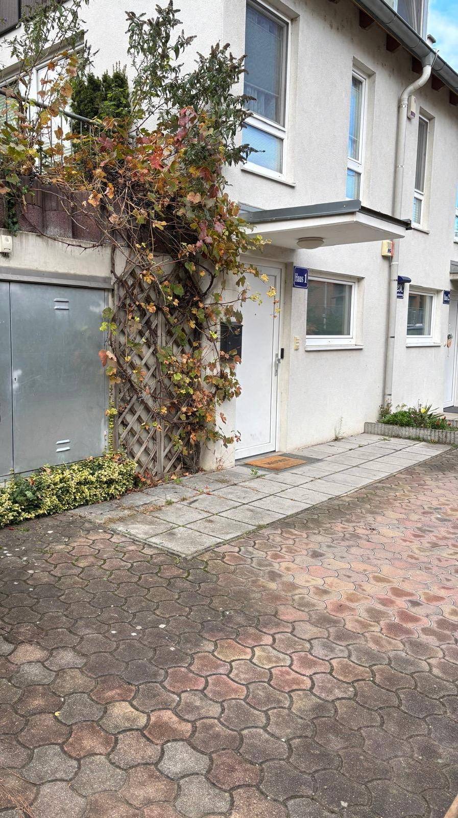 Eingangsbereich eines Reihenhauses mit Kletterpflanzen und gepflastertem Vorplatz.