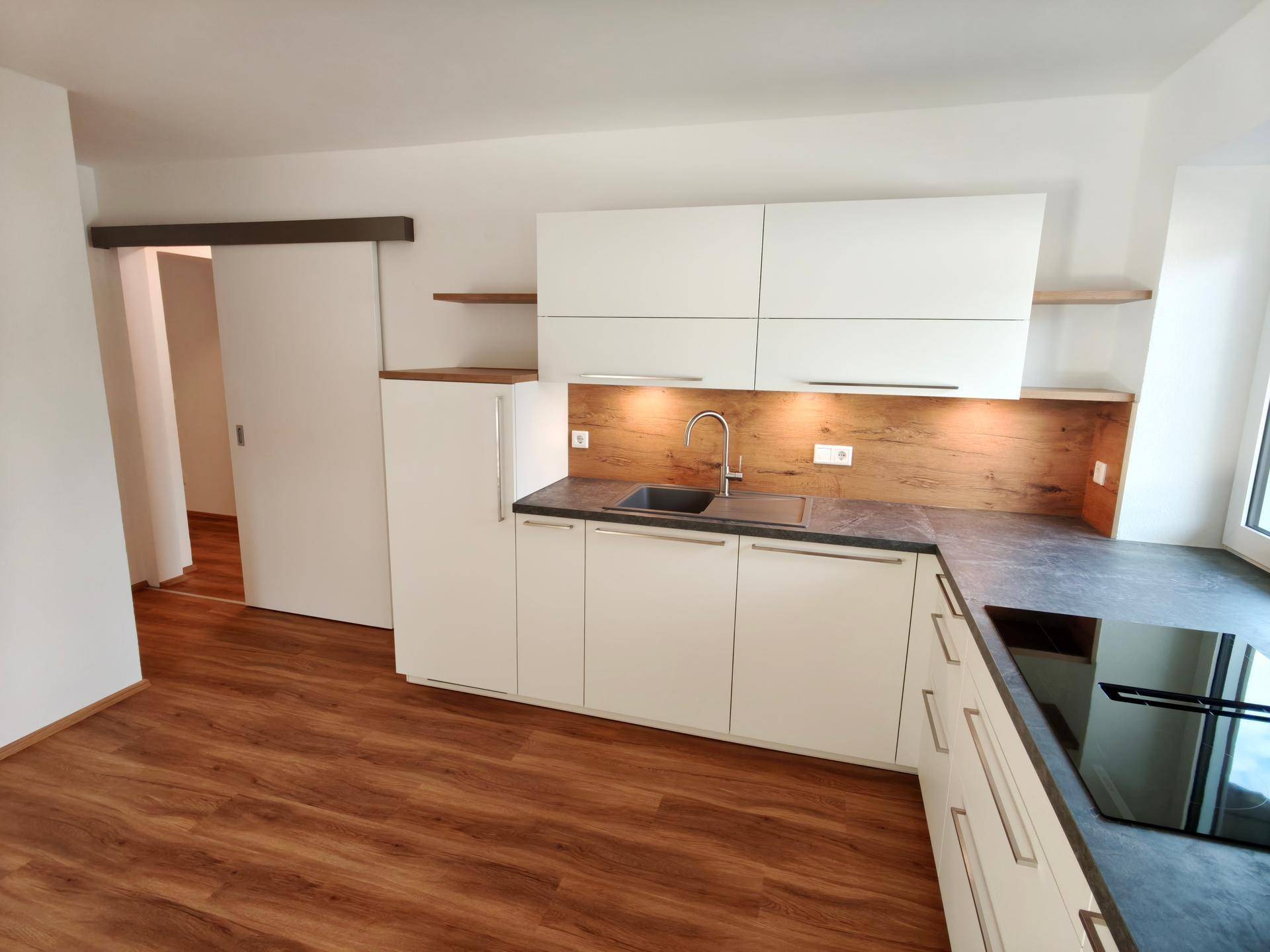 Helle L-förmige Küche mit weißen Schränken, Holzrückwand und Zugang zu einem weiteren Raum.
