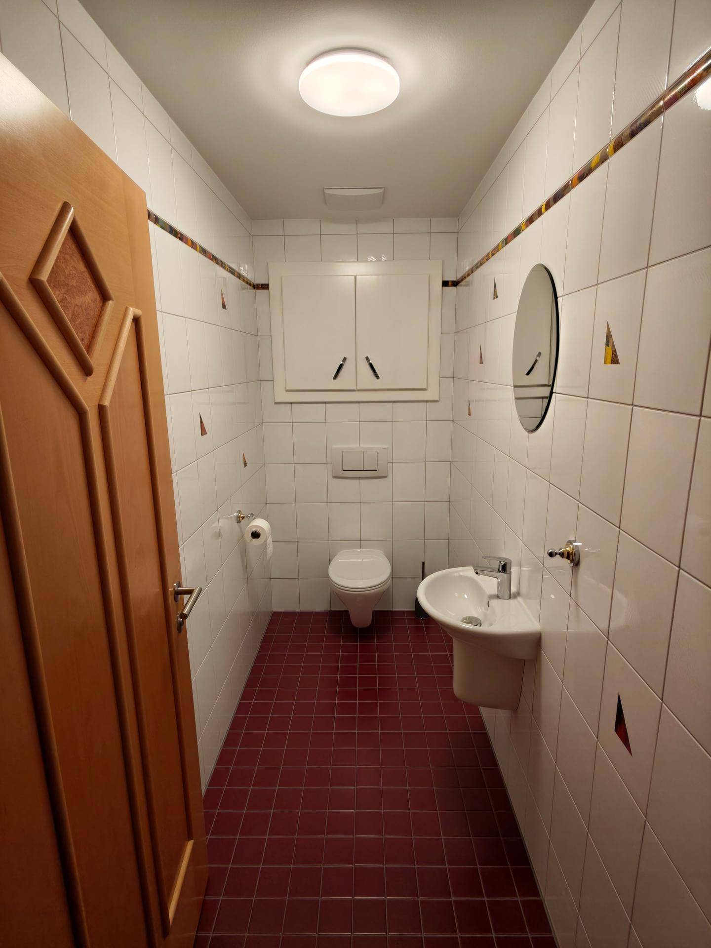 Funktionales Gäste-WC mit weißen Wandfliesen, roten Bodenfliesen, einem Waschbecken und praktischem Wandschrank.