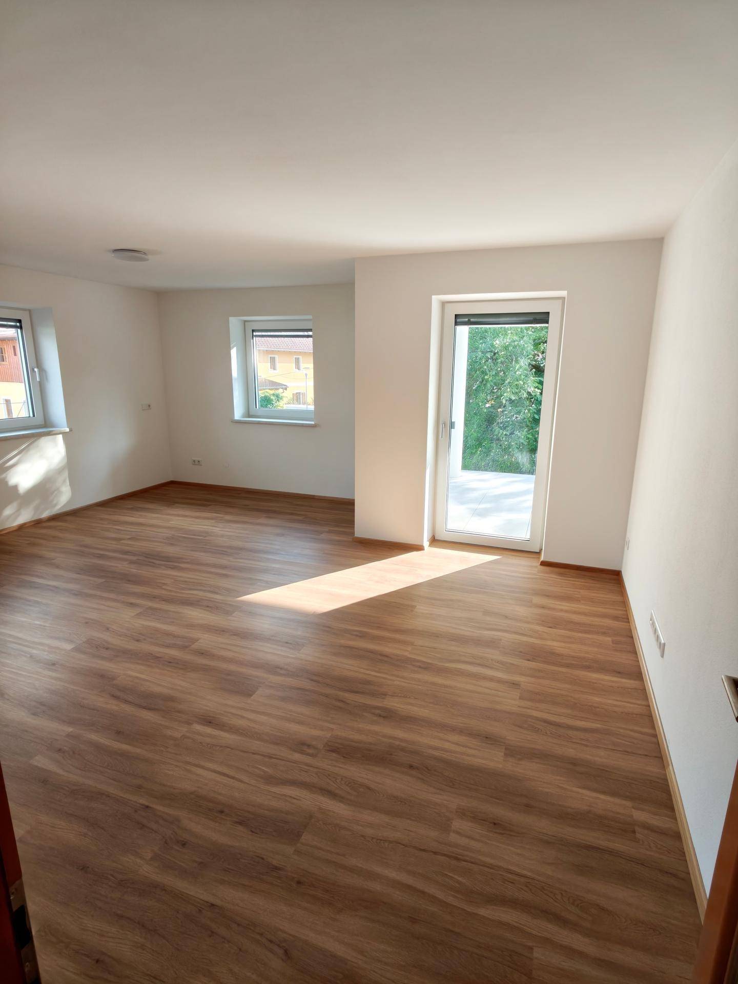 Geräumiger, heller Raum mit Laminatboden, großen Fenstern und Zugang zur Terrasse.