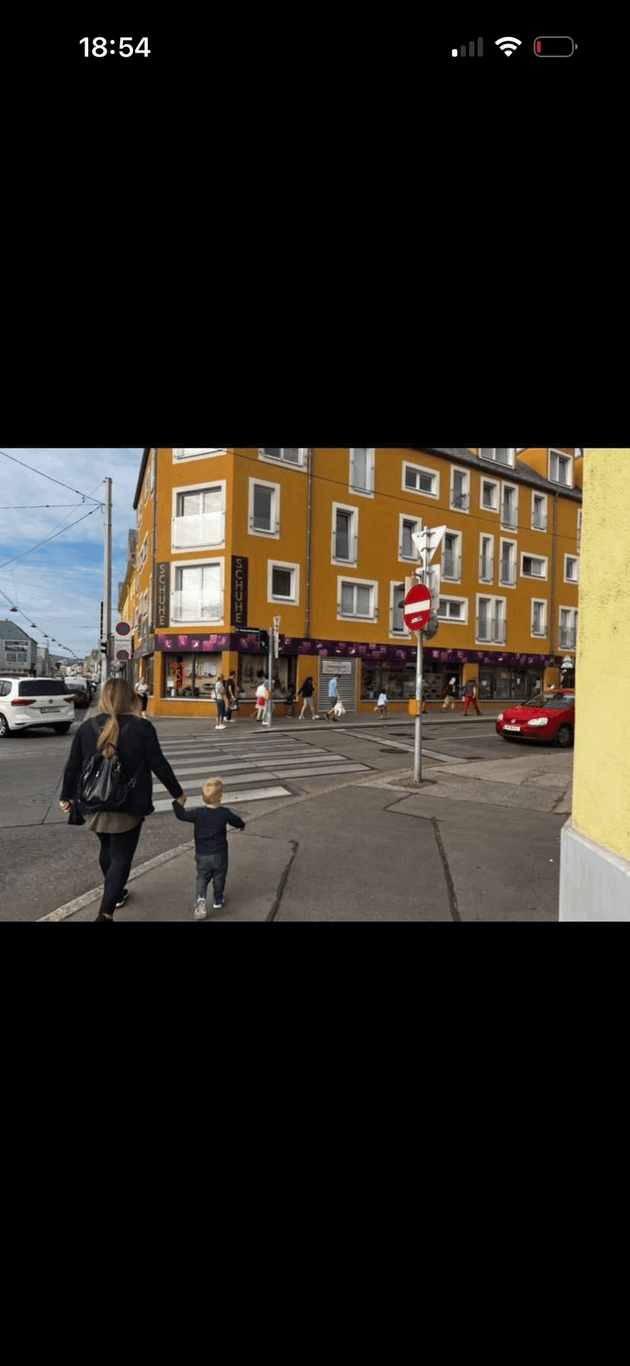 Belebte Straßenszene mit einem gelben Gebäude, Passanten und einem roten Auto im Hintergrund.