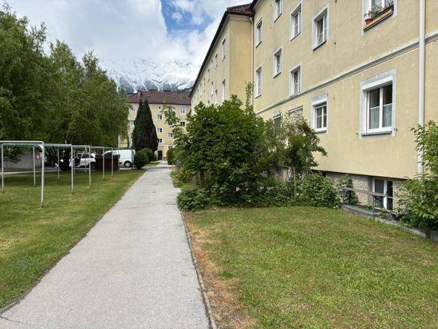 Außenansicht eines Mehrfamilienhauses mit Grünflächen und Bergen im Hintergrund.