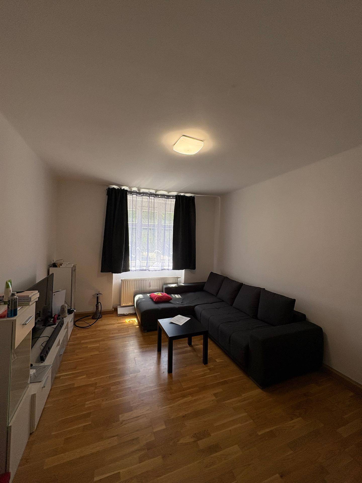 Wohnzimmer mit dunklem Sofa, Holzboden und Fenster mit schwarzen Vorhängen.