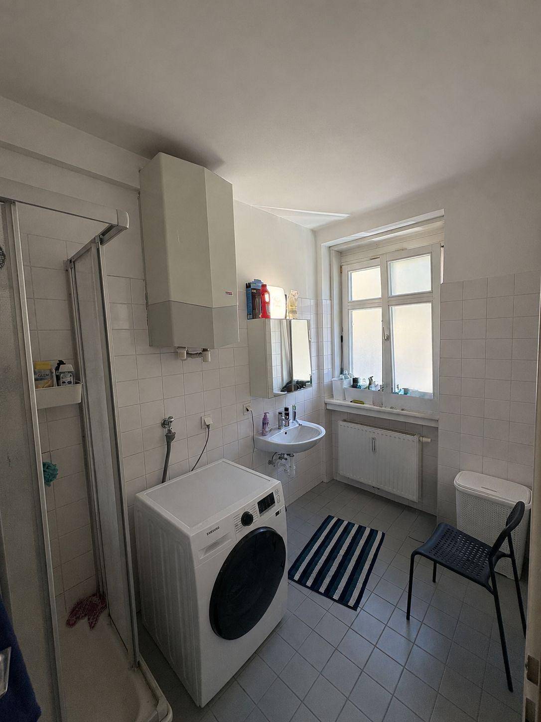 Badezimmer mit Dusche, Waschbecken, Waschmaschine und Fenster, gefliest.