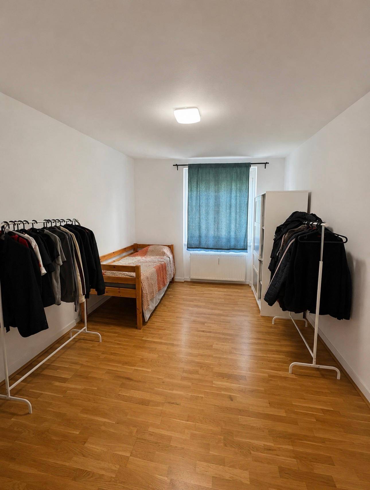 Schlafzimmer mit Holzboden, Einzelbett und Kleiderständern, gut beleuchtet durch ein Fenster.