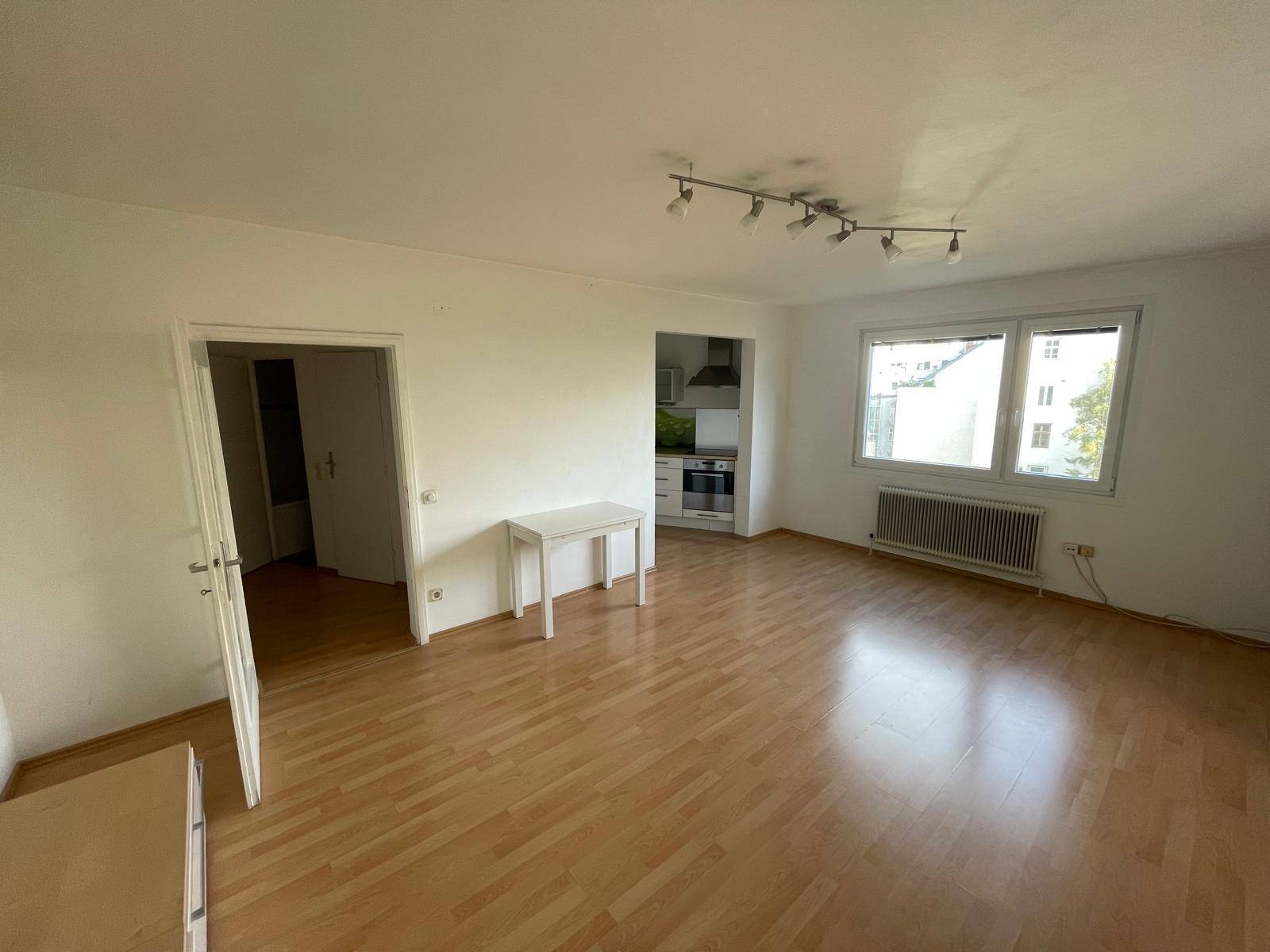 Geräumiges, leeres Wohnzimmer mit hellem Parkettboden, großen Fenstern und Zugang zur Küche.