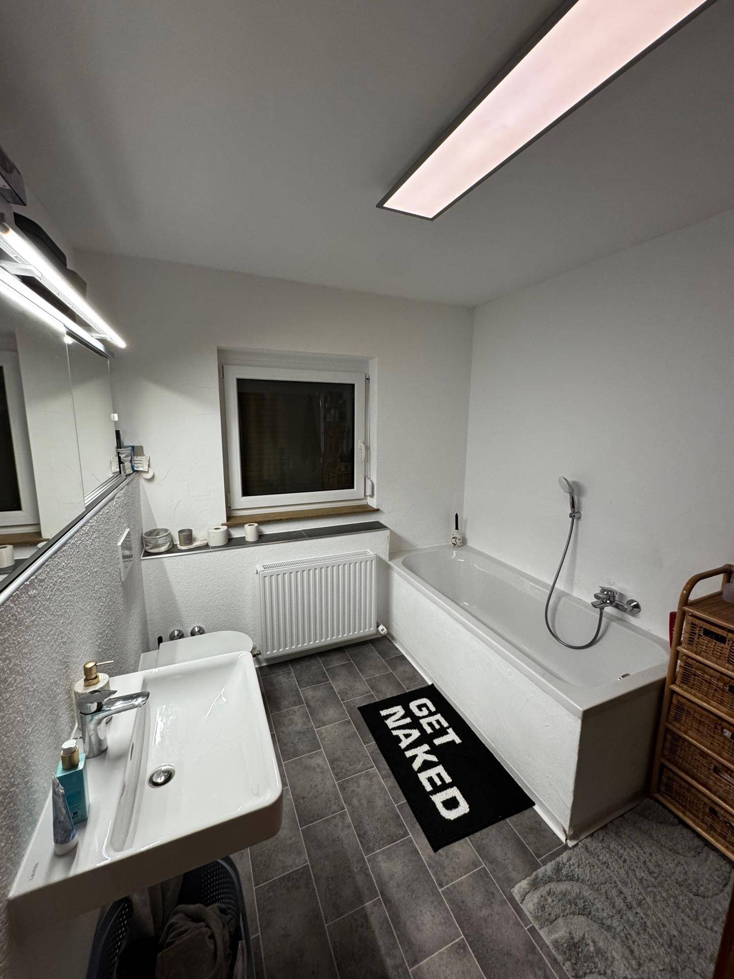 Badezimmer mit Badewanne, Toilette, Waschbecken und einem Fenster, ausgestattet mit grauen Fliesen.