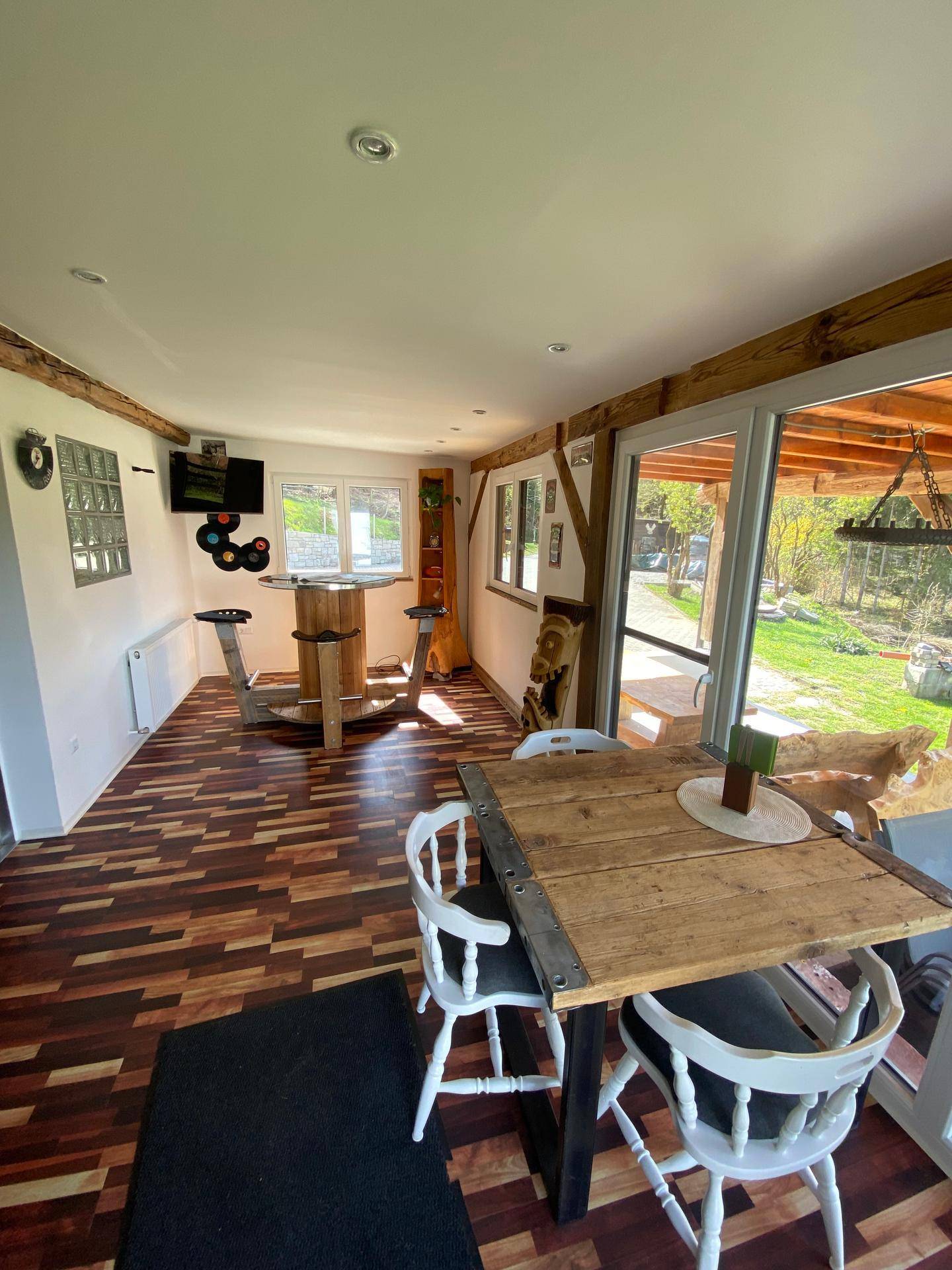 Ess- und Loungebereich mit Holztischen, Stühlen und großen Fenstern mit Blick auf eine Grünfläche.