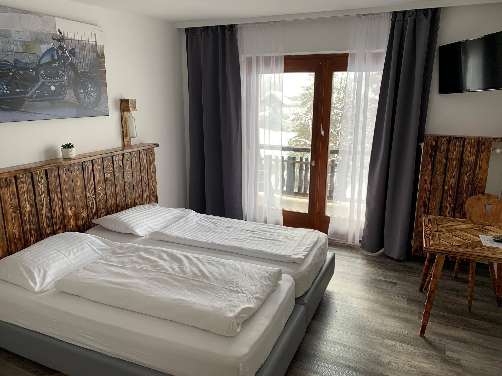 Schlafzimmer mit zwei Betten, rustikalem Holz-Kopfteil, kleinem Holztisch und einer Balkontür.
