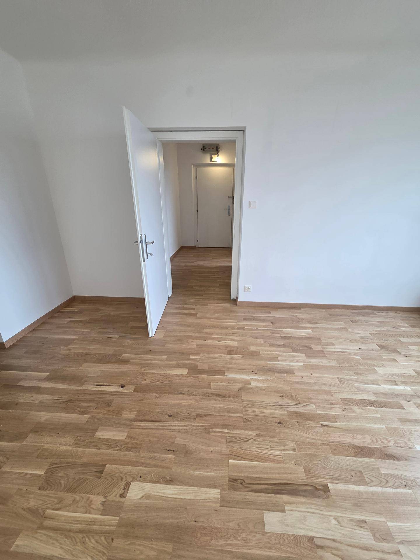 Leerer Raum mit hellem Holzparkett und weiß gestrichenen Wänden, mit Zugang zum Flur.