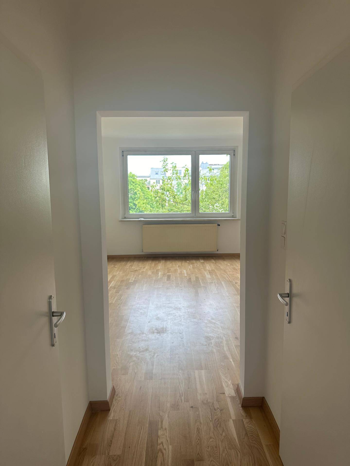 Heller Raum mit Holzparkett und großem Fenster, das einen grünen Ausblick bietet.