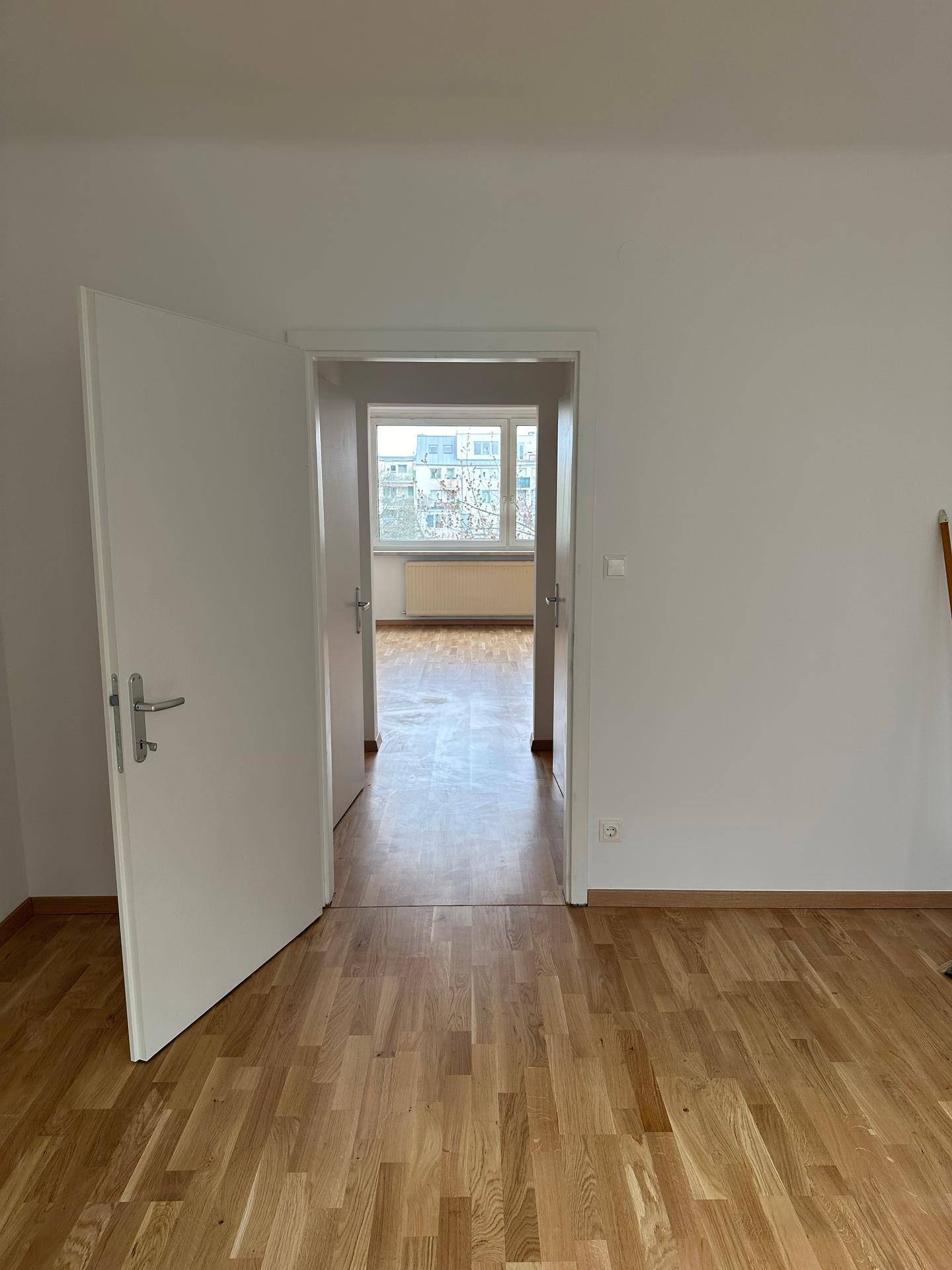 Heller Raum mit Holzparkett und Blick durch eine Tür in ein weiteres Zimmer mit Fenster.