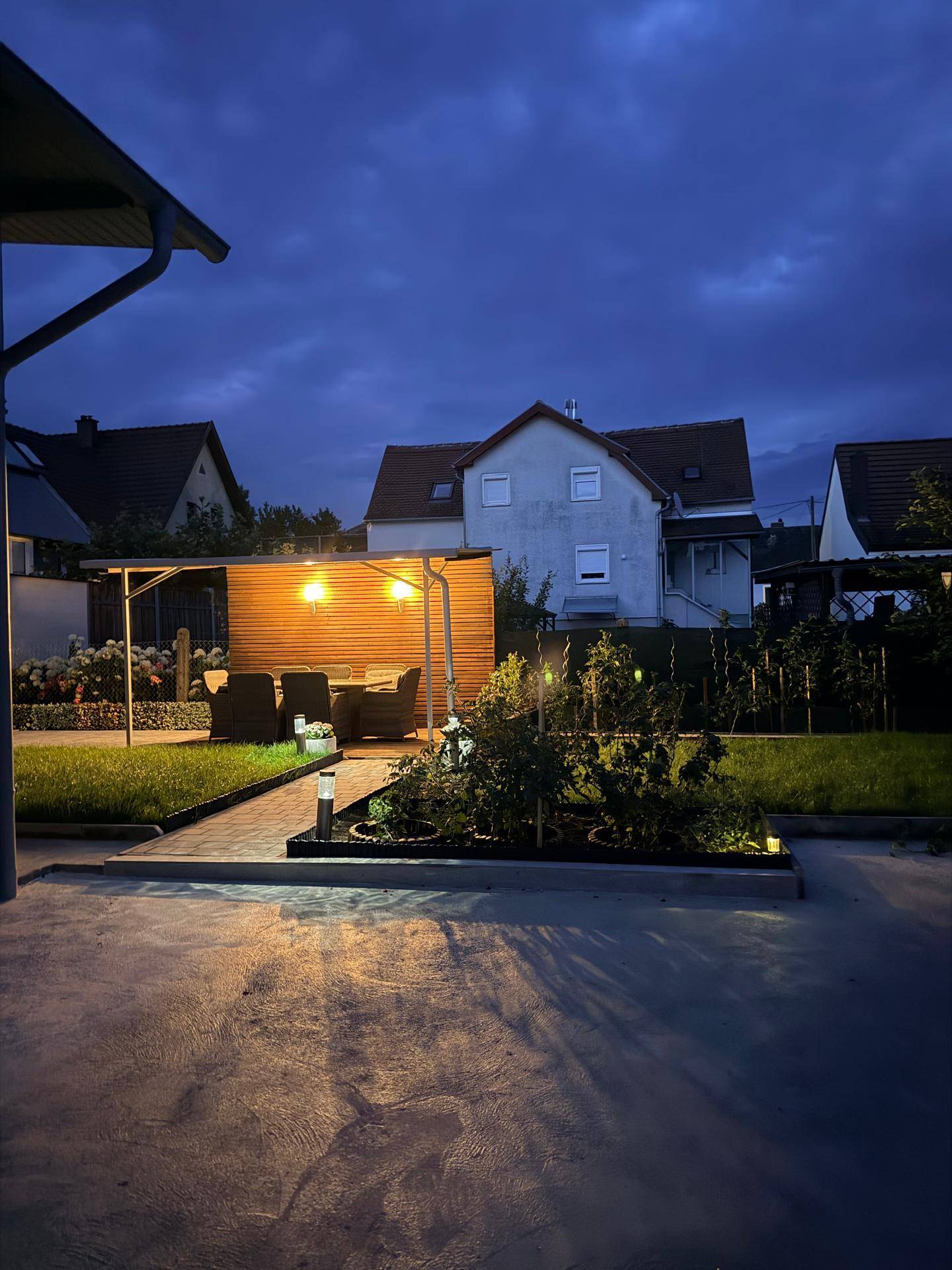 Abendlicher Blick auf einen beleuchteten Garten mit überdachtem Sitzbereich und Wohnhäusern im Hintergrund.