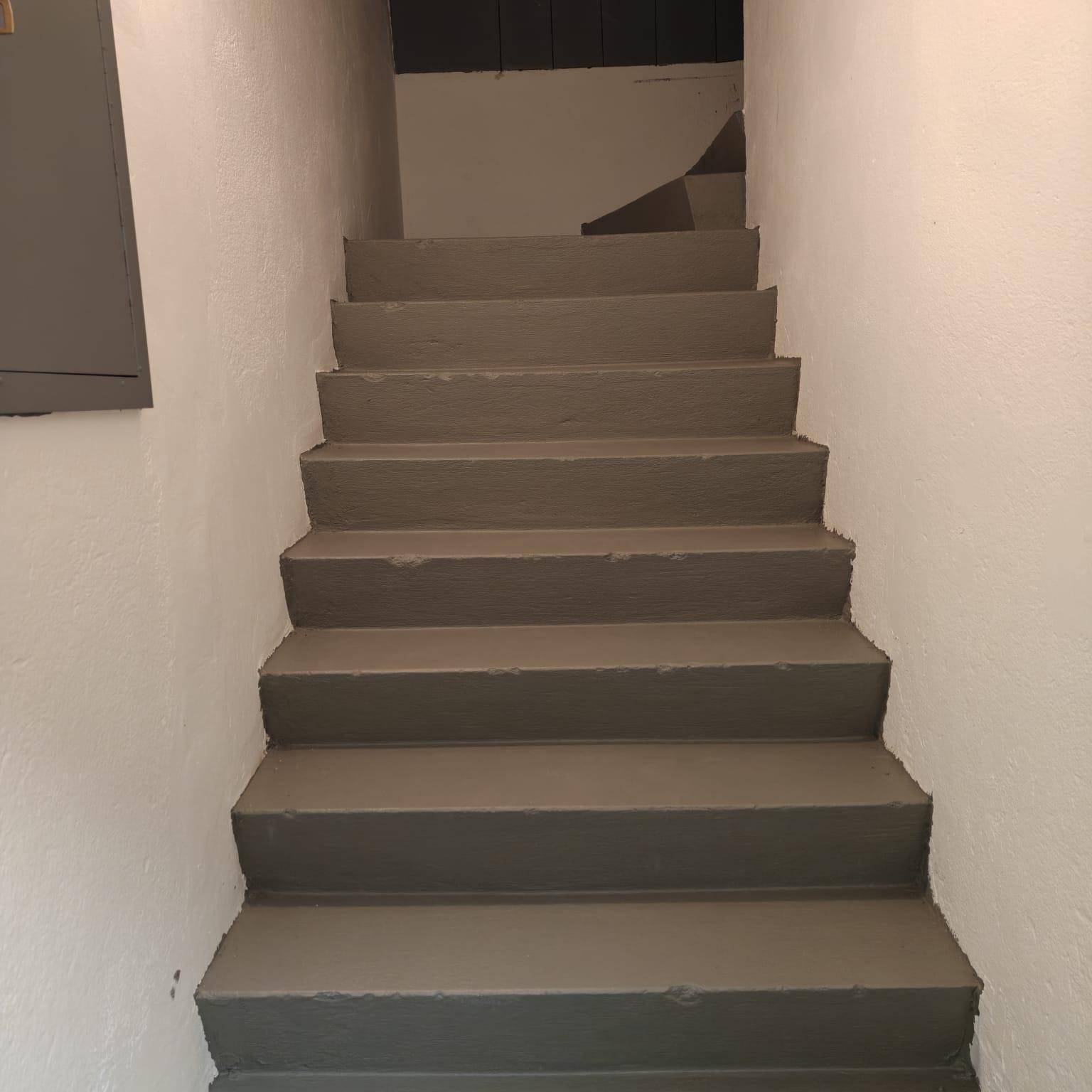 Einfache Treppe mit grauen Stufen und weißen Wänden, die in ein Obergeschoss führt.