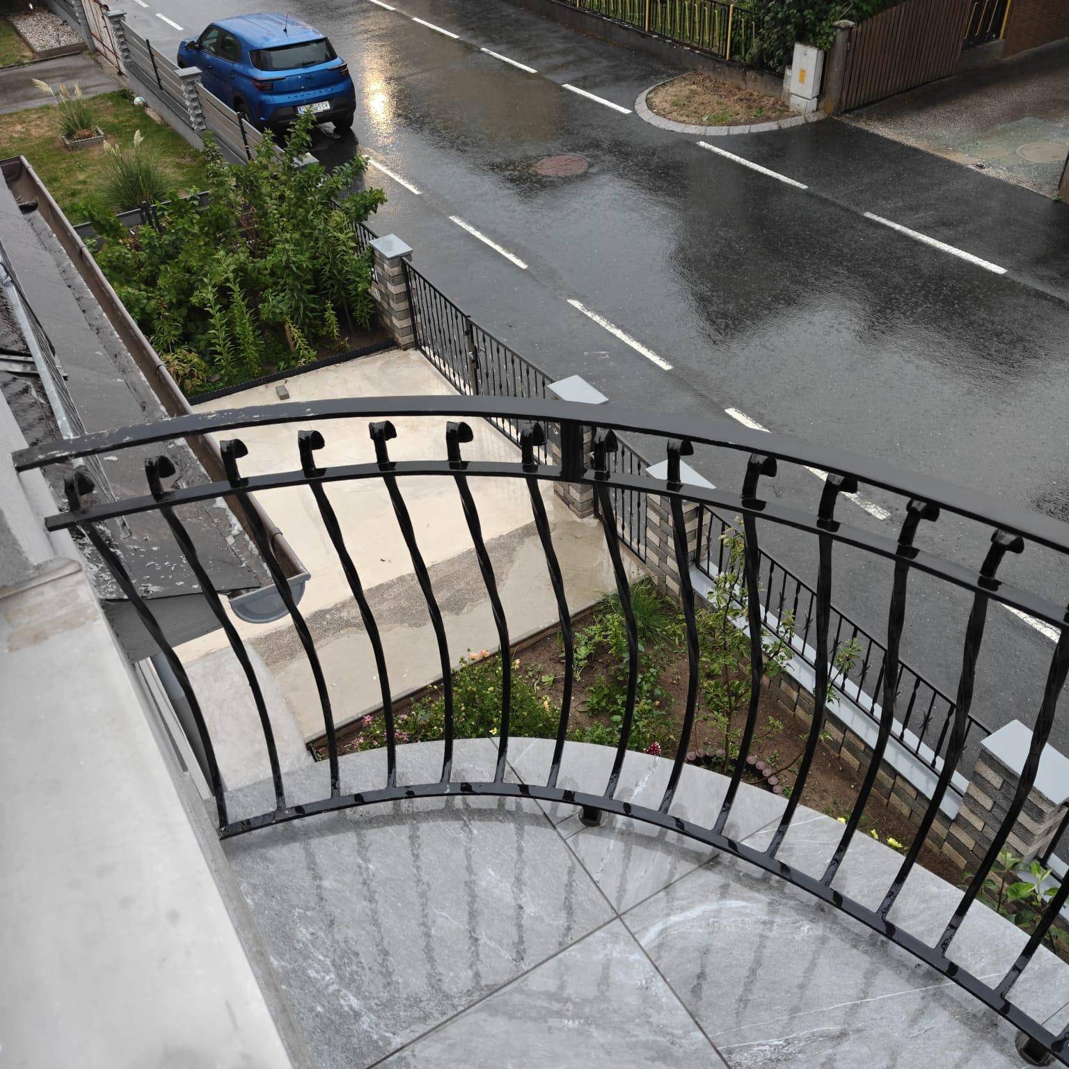 Blick von einem Balkon mit schmiedeeisernem Geländer auf eine nasse Straße und einen Vorgarten.