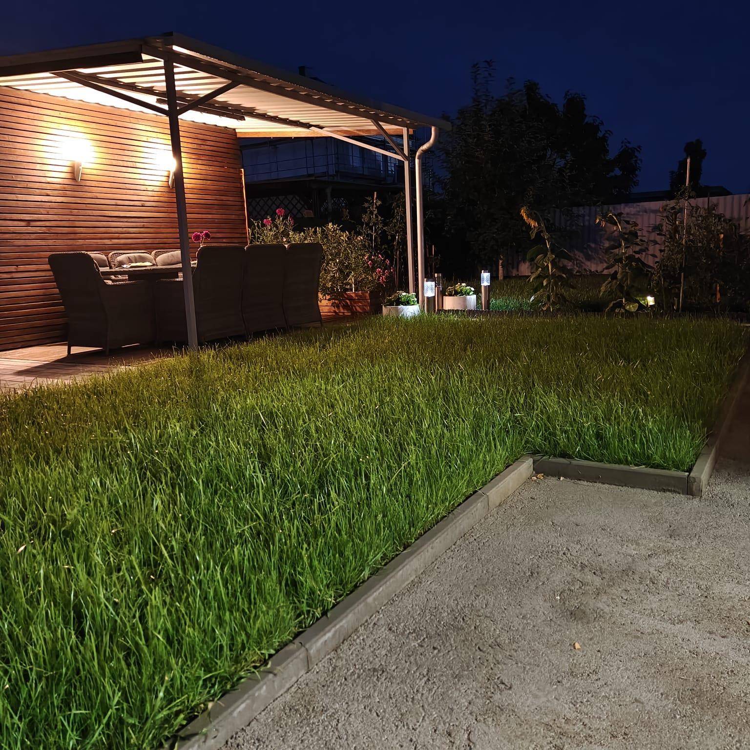 Große Rasenfläche im Garten bei Nacht, mit einem beleuchteten, überdachten Sitzbereich im Hintergrund.