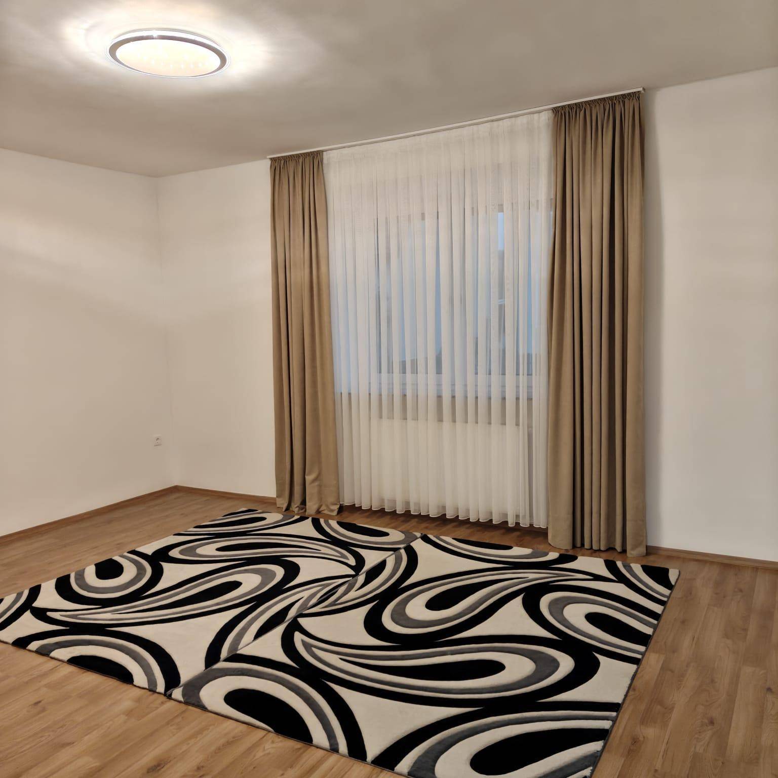 Helles Zimmer mit Laminatboden, großen Fenstern und beigen Vorhängen.