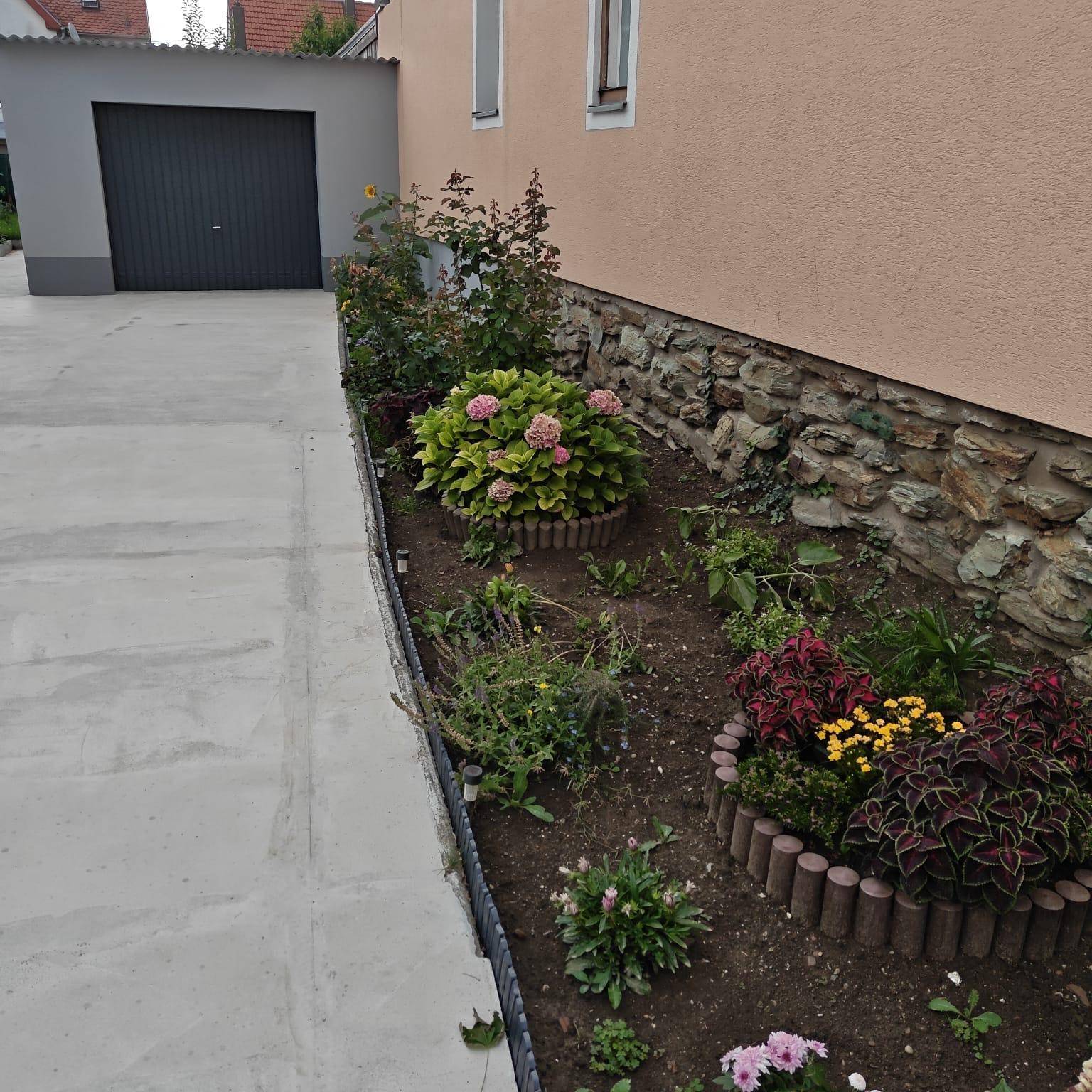 Schmaler Gartenstreifen mit verschiedenen Pflanzen und einem gepflasterten Bereich neben der Garage.