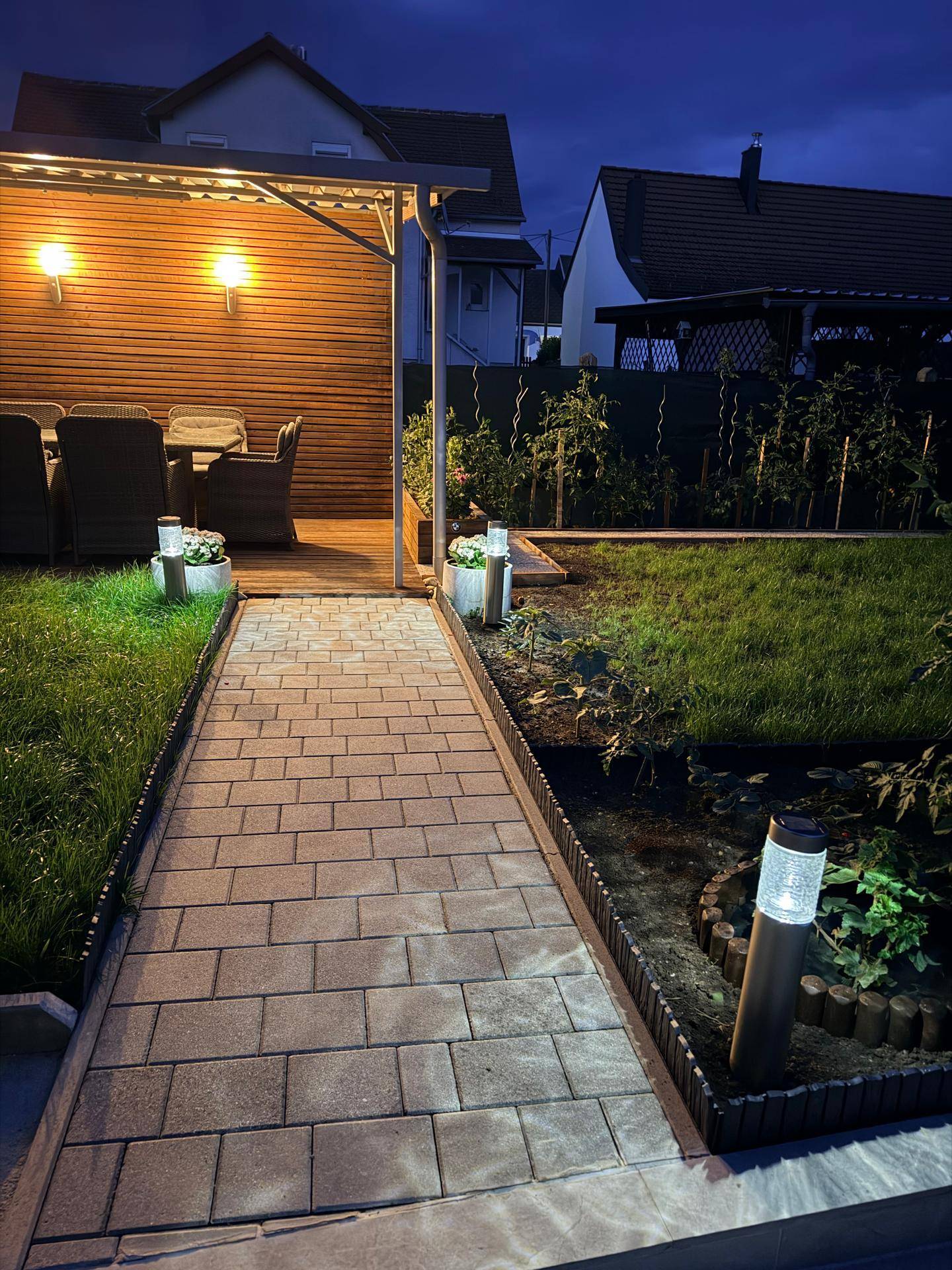 Beleuchteter Gartenweg führt zu einem gemütlichen, überdachten Sitzbereich, umgeben von Grünflächen.