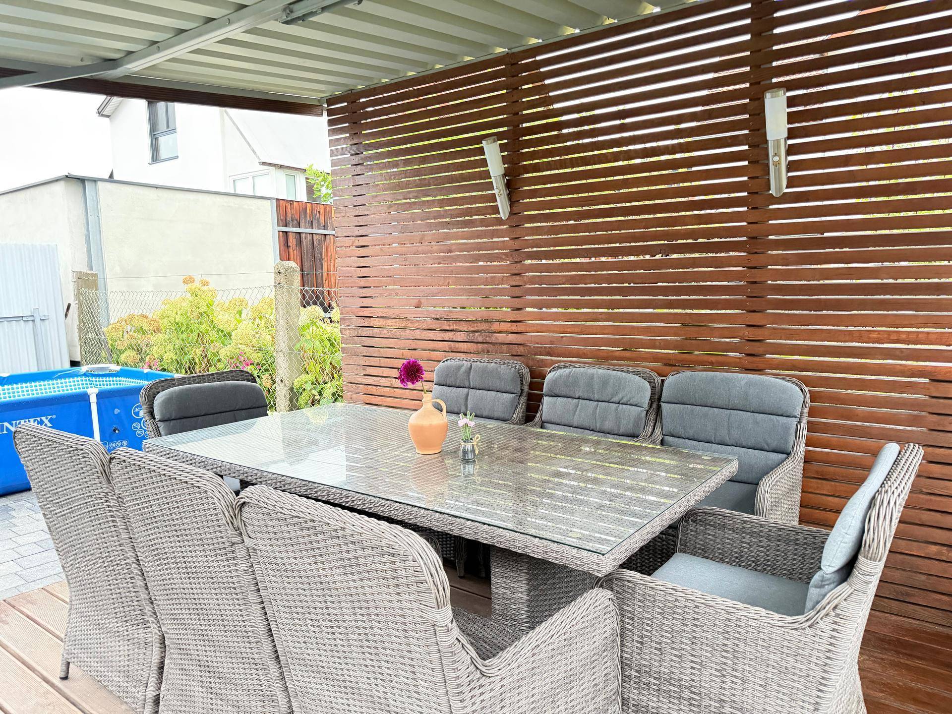 Überdachte Terrasse mit Esstisch und Korbstühlen, umgeben von einer Holzwand und Blick auf den Garten.