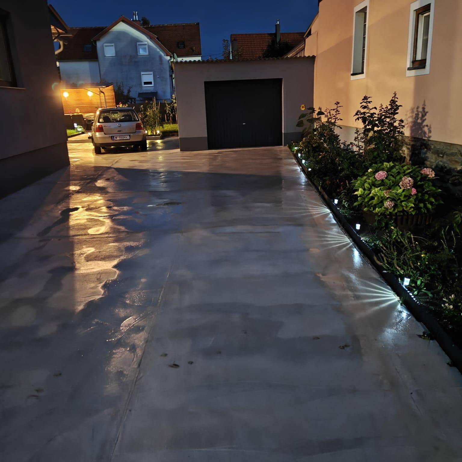 Beleuchtete Einfahrt führt zu einer Garage, gesäumt von bepflanzten Beeten mit Bodenleuchten.