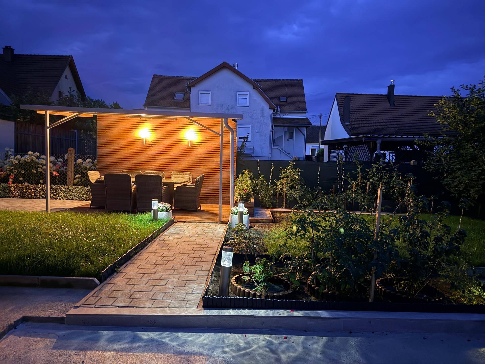 Ruhiger Abend im Garten mit beleuchteter Terrasse und gepflegten Beeten vor den Nachbarhäusern.