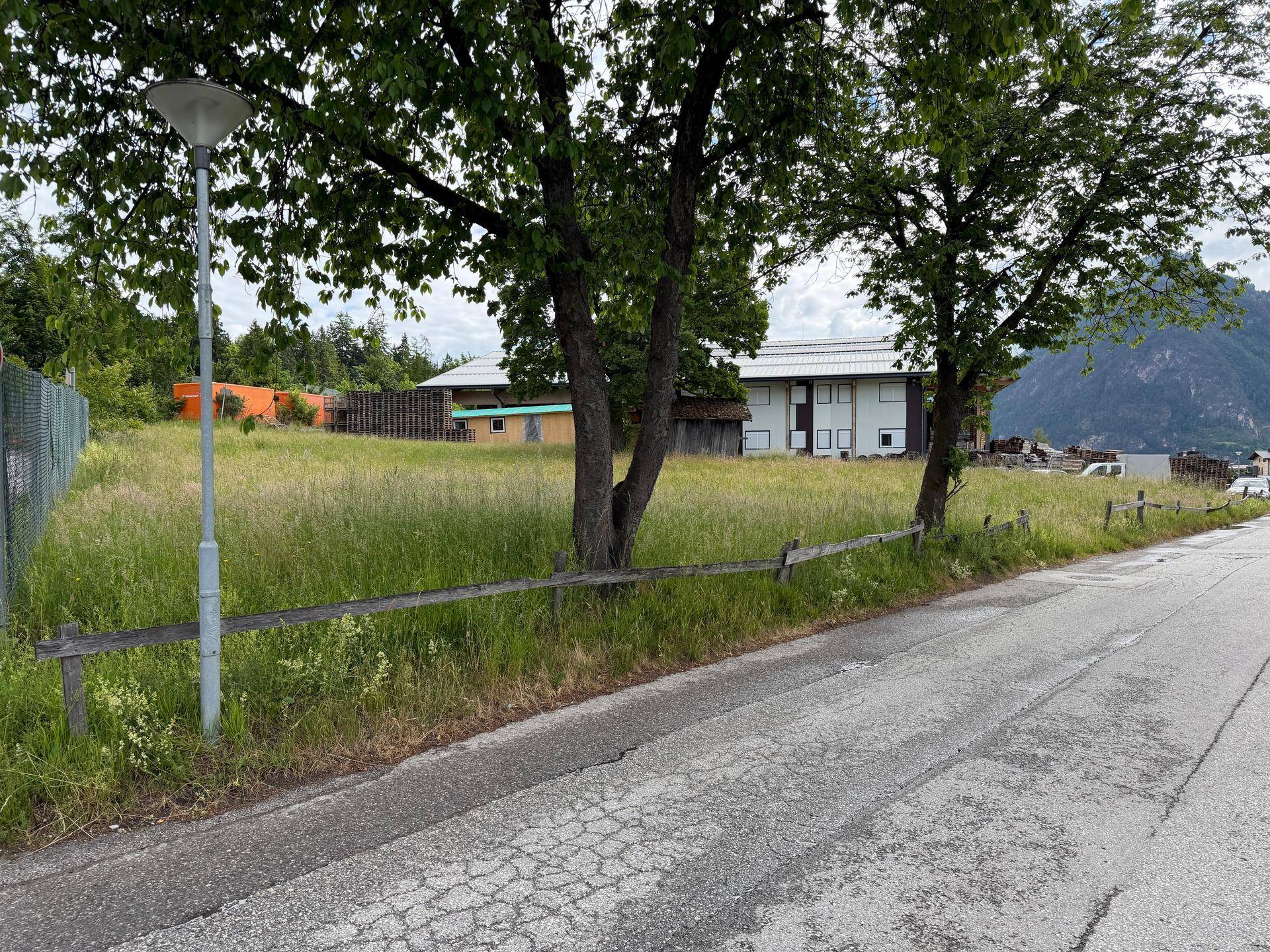 Blick von der Straße auf ein großes, grünes Grundstück mit Bäumen und Gebäuden im Hintergrund, umgeben von Bergen.