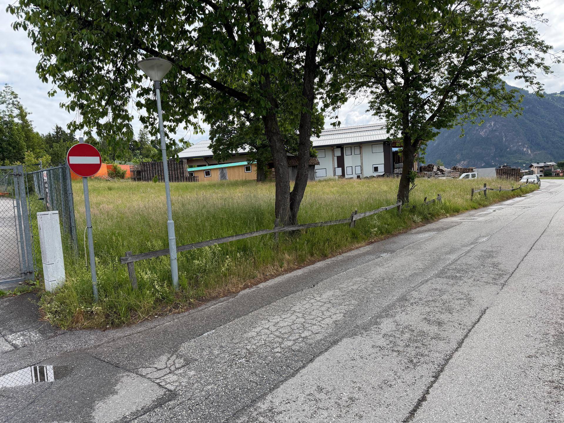 Ansicht eines grünen Grundstücks von der Straße aus, mit Bäumen, einem Verkehrsschild und Bergen im Hintergrund.