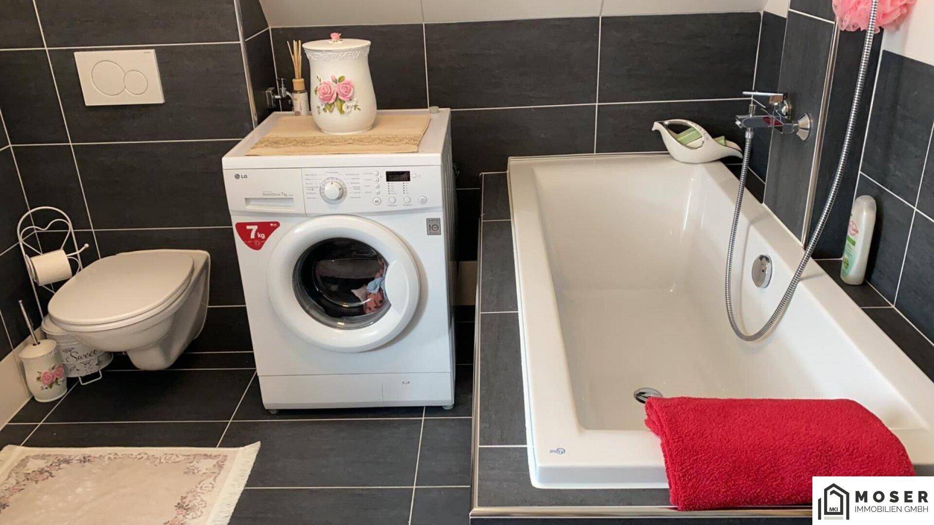 Badezimmer mit Badewanne, Toilette und Waschmaschine, ausgestattet mit dunklen Fliesen.