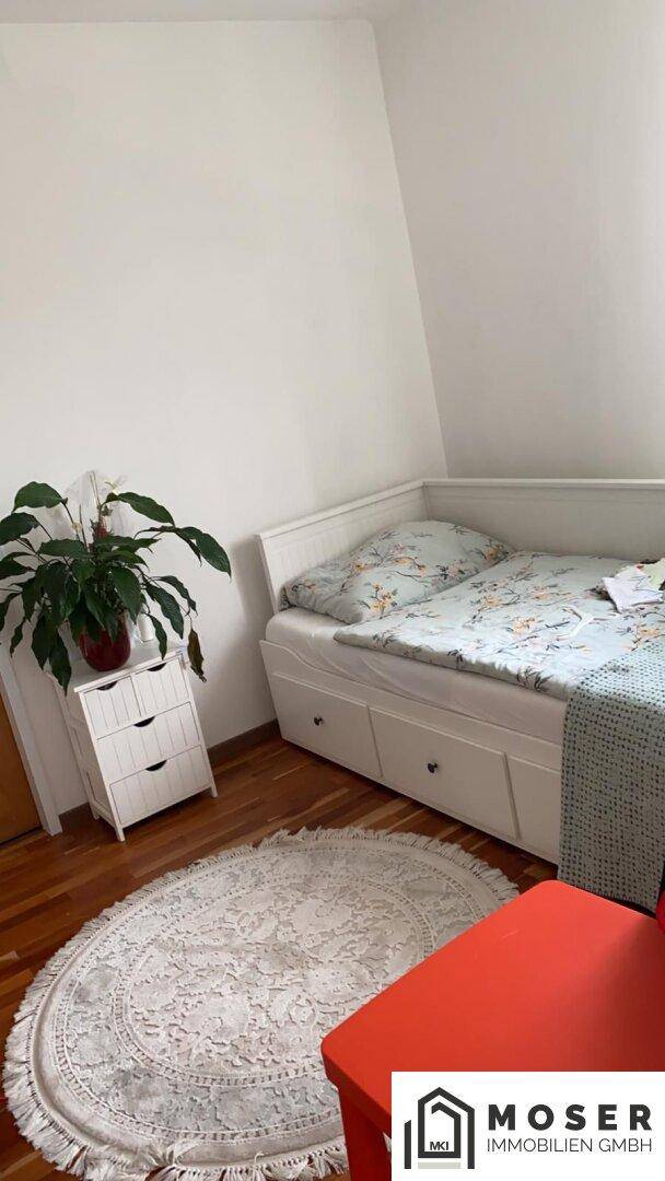 Helles Schlafzimmer mit Einzelbett, Nachttisch und einem runden Teppich auf Parkettboden.