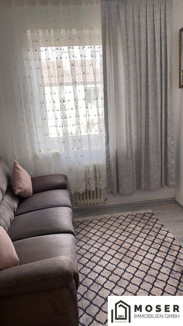 Gemütliches Wohnzimmer mit Sofa und gemustertem Teppich vor einem Fenster.