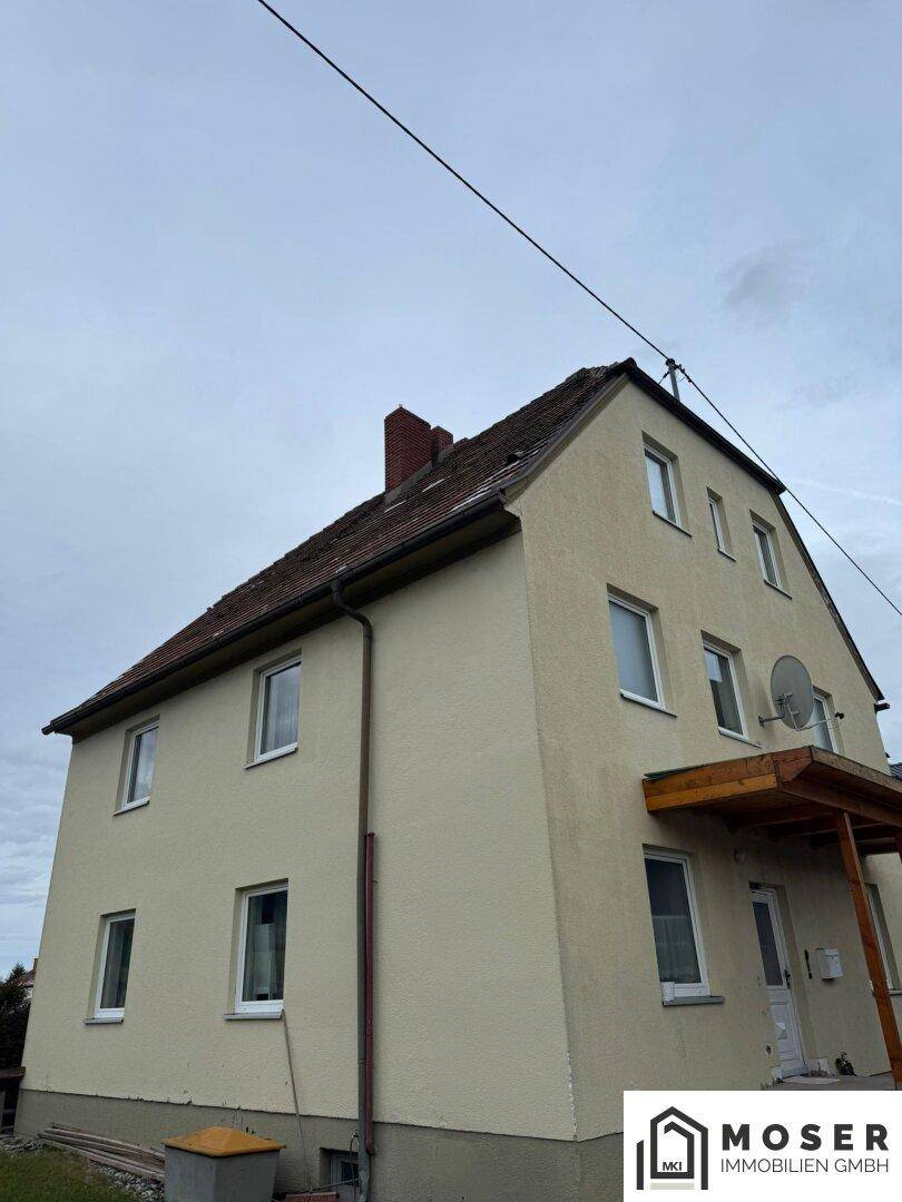 Seitenansicht eines Hauses mit beiger Fassade, Satteldach und einem überdachten Eingangsbereich.