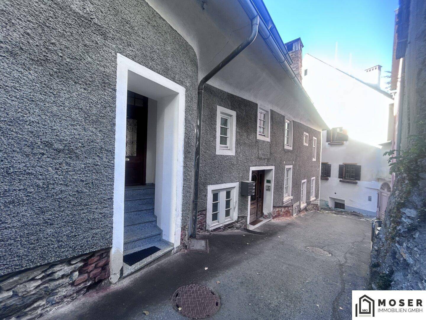 Eingangsbereich eines Gebäudes in einer engen Gasse mit grauer Fassade und Steinstufen.