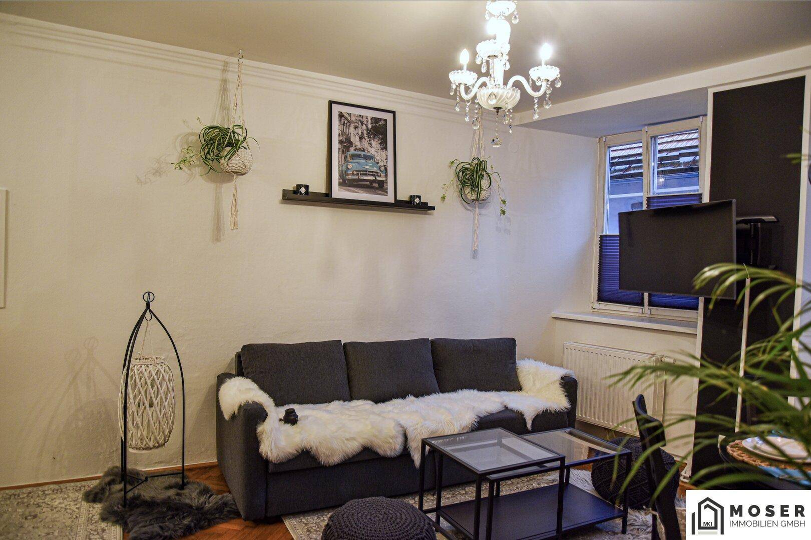 Gemütliches Wohnzimmer mit dunklem Sofa, Fellteppichen und einem Kronleuchter, dekoriert mit Pflanzen.