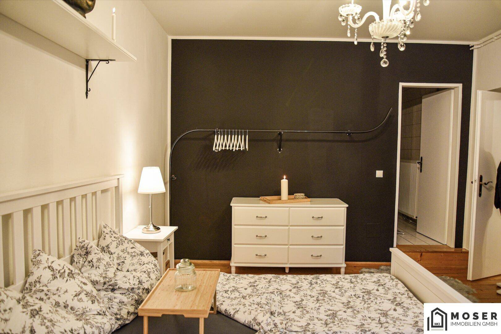 Schlafzimmer mit weißem Bett, dunkler Akzentwand, Kommode und einem eleganten Kronleuchter.