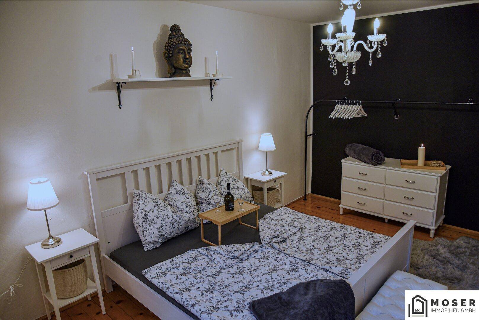 Schlafzimmer mit weißem Bett, dunkler Akzentwand, Kommode und einem eleganten Kronleuchter.