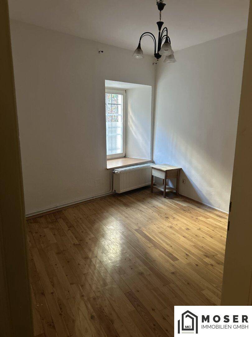 Leeres Zimmer mit Holzboden, weiß gestrichenen Wänden und einem Fenster mit Heizkörper.