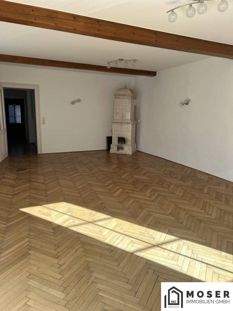 Geräumiges Wohnzimmer mit Fischgrätparkett, Kachelofen und Holzbalken an der Decke.