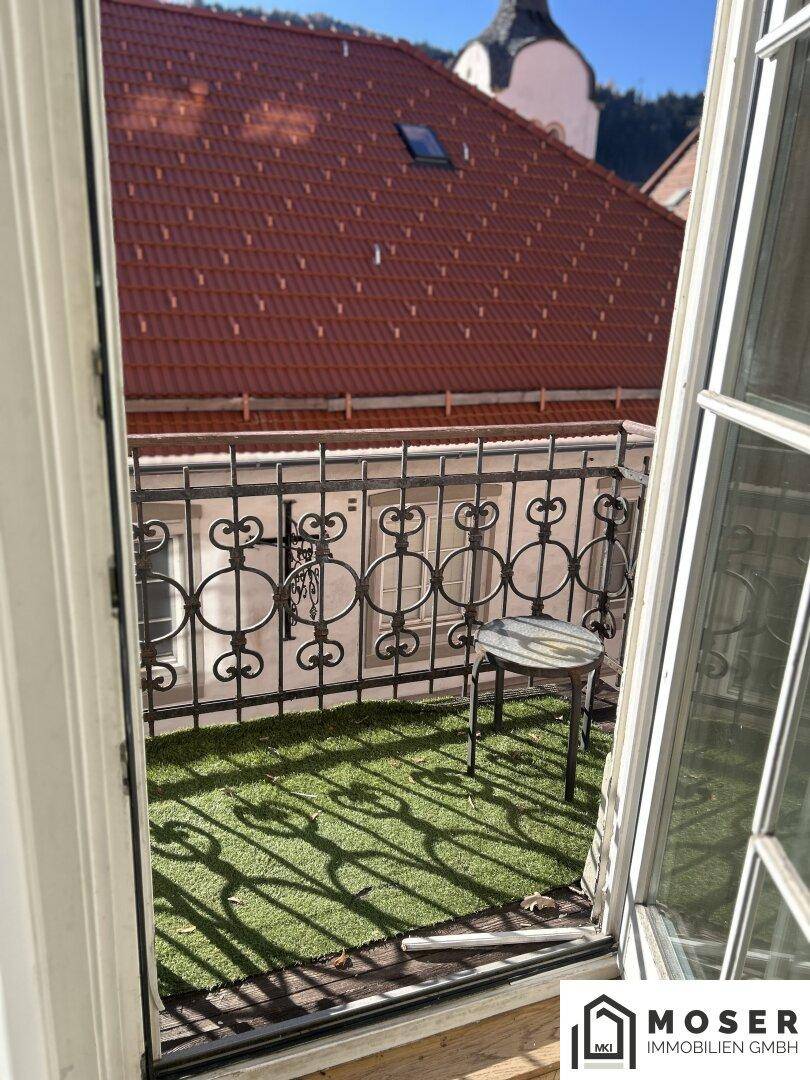 Kleiner Balkon mit Kunstrasen, schmiedeeisernem Geländer und Blick auf die Dächer.
