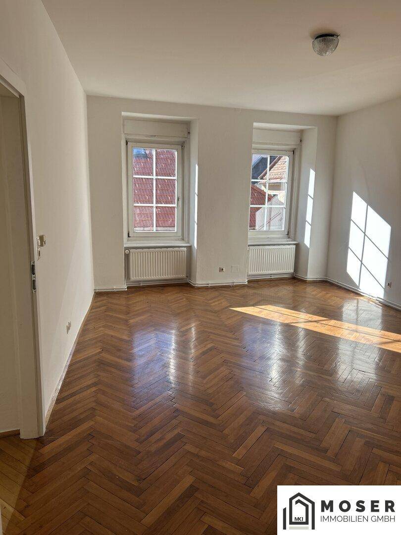 Helles Zimmer mit Fischgrätparkett und zwei Fenstern, die viel Tageslicht hereinlassen.