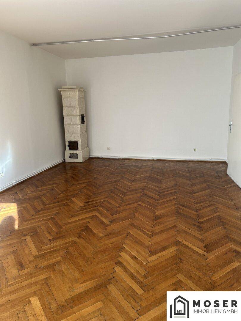 Geräumiges Zimmer mit Fischgrätparkett, weiß gestrichenen Wänden und einem Kachelofen.